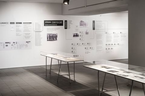 Installation. Two long display cases filled with papers and images are in a gallery space lined with an exhibit of text and images on the wall opposite us.
