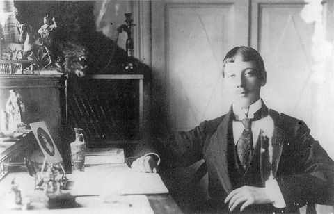 Black and white photograph. A man with pale skin sitting next to a desk, to our right, wears a high-collared white shirt, long tie, vest, and jacket. His hair is parted down the middle and bangs cut straight across, high on his forehead.