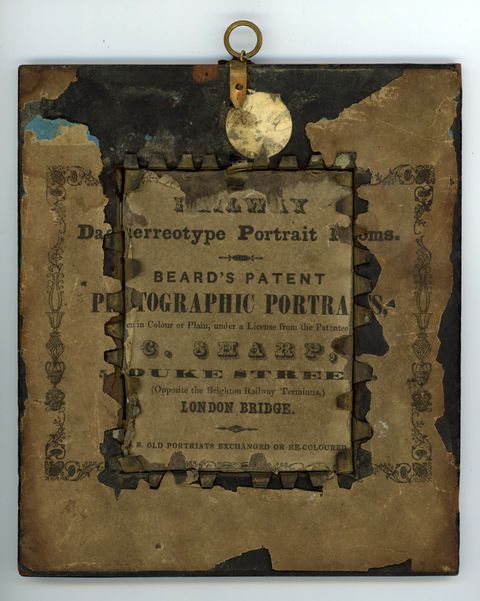 Label. A printed label is affixed to the back of a daguerreotype. The paper is chipped and discoloured, but some words and phrases can be made out. It reads as follows: “Daguerreotype Portrait Rooms Beard’s Patent Photograph Portraits...in Colour or Plain, under a License from the Patentee C. Sharp, Duke Street (Opposite the Brighton Railway Terminus,) London Bridge. Old Portraits Exchanged or Recoloured”.