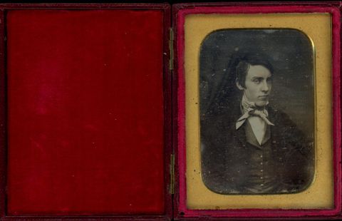 Daguerreotype in an open case. A pale-skinned, clean-shaven young man’s face is angled to our right, but he looks at us under heavy brows, his chin slightly tucked. The rectangular mat has curved edges. The crimson-red case is flipped open to our left.