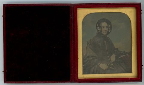 Daguerreotype in an open case. A pale-skinned woman wearing a bonnet and black dress has been hand-coloured to add rosy cheeks and a blue background. The photograph is in a gold frame in a red case, which opens like a book. The cover is lined with black fabric.