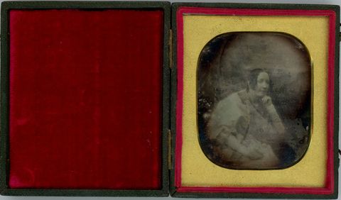 Daguerreotype. A pale-skinned woman with curls framing her long face looks off to our right, chin resting in one hand. The background is difficult to make out due to some abrading or discoloration. The gold mat is rounded at the corner.