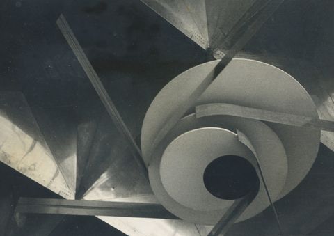 Installation photograph. We look up at an abstract sculpture hanging from deep, angular ceiling coffers. The sculpture has four discs in descending sizes. On each, one or two metal strips metal are affixed to the narrow edges and extend off at tangents. The photograph is black and white. 