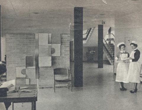 Black and white photograph. Two uniformed nurses stand to one side of an open space with staircases at the back corner, to our right. To our left, square and rectangular panels create a cascading form against a brick wall.