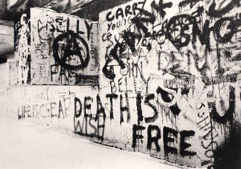 Architecture. A concrete wall is covered with graffiti in this black and white photograph. Some painted words and phrases read “Life is Cheap Death is Free”, “Wish”, “Belly,” and “Young”. A letter “A” within a circle is to our left.