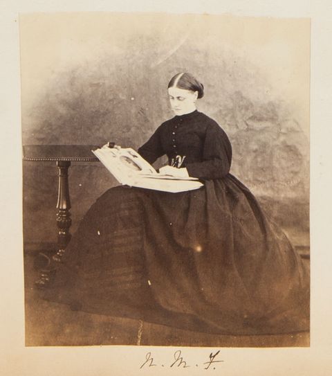 Black and white photograph. A pale-skinned woman wearing a long, puffy-sleeved dress with a full, wide skirt sits looking at an album. Her dark hair is parted down the middle and pulled back at the nape of her neck. A side table sits next to her right hand, to our left.