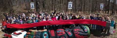 Dozens of people stand holding or behind a long strip of cloth in front of a line of leafless trees. A totem pole painted in red, black, blue, white, and green lies on the ground in front of the group. Several people in the crowd hold up fists.
