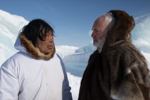 Video still. Two men stand near us. To our left, a man wears a white parka with a fur-lined hood. He has dark skin, black hair, and a grey moustache. He talks to a pale-skinned man, to our right. The latter has a white beard and hair, and wears a fur parka with the hood up. They stand in front of a snowy expanse.