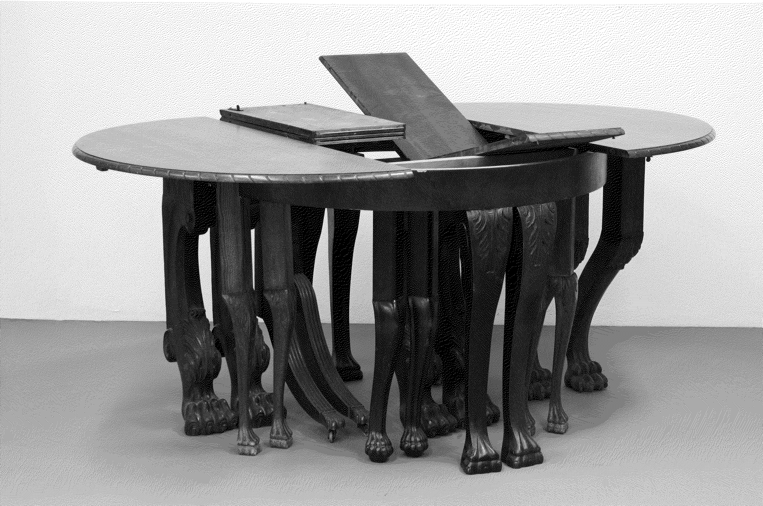 Sculpture. The two halves of a round dining room table have been pulled apart. The leaves that would expand its length sit, partially unfolded or totally folded up, in the gap. Ten pairs of dining room legs, all with clawed feet, hold the table up.